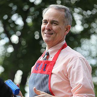 Photo of John 麦克维恩 smiling wearing a striped apron and holding a camera and blue cup